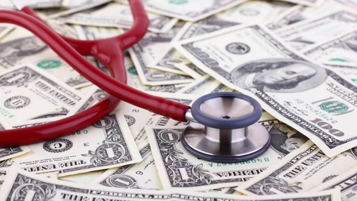 A red stethoscope resting on a spread of scattered US dollar bills, with the chest piece visible at the centre