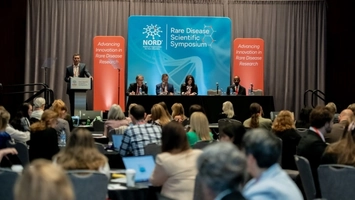 Panel of four speakers at a conference table addressing a seated audience in a conference room