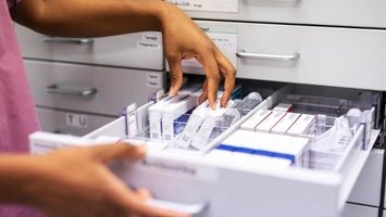 Hands sorting packaged medications in a pharmacy drawer with labelled drug boxes arranged in compartments